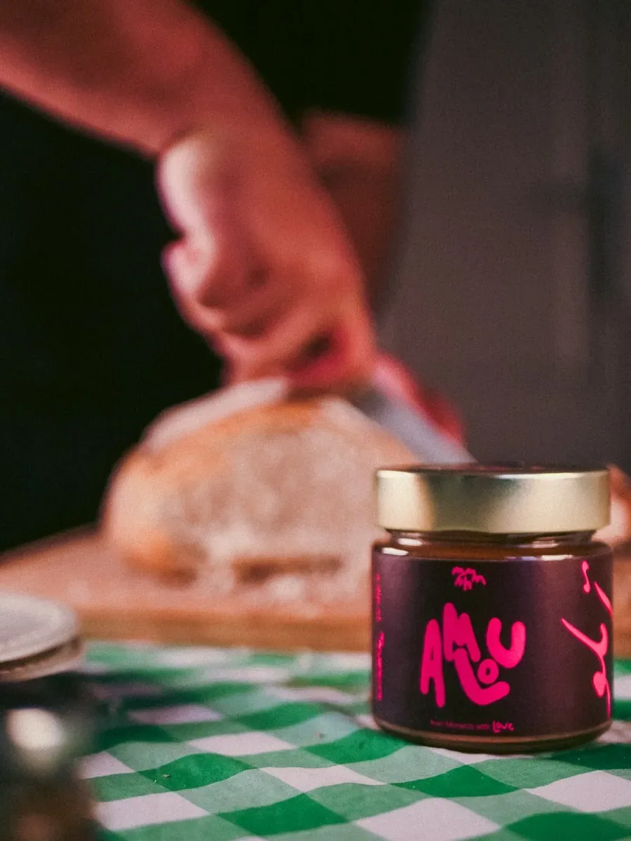Jar of MMMHH amlou on a green gingham tablecloth while bread is sliced in the background