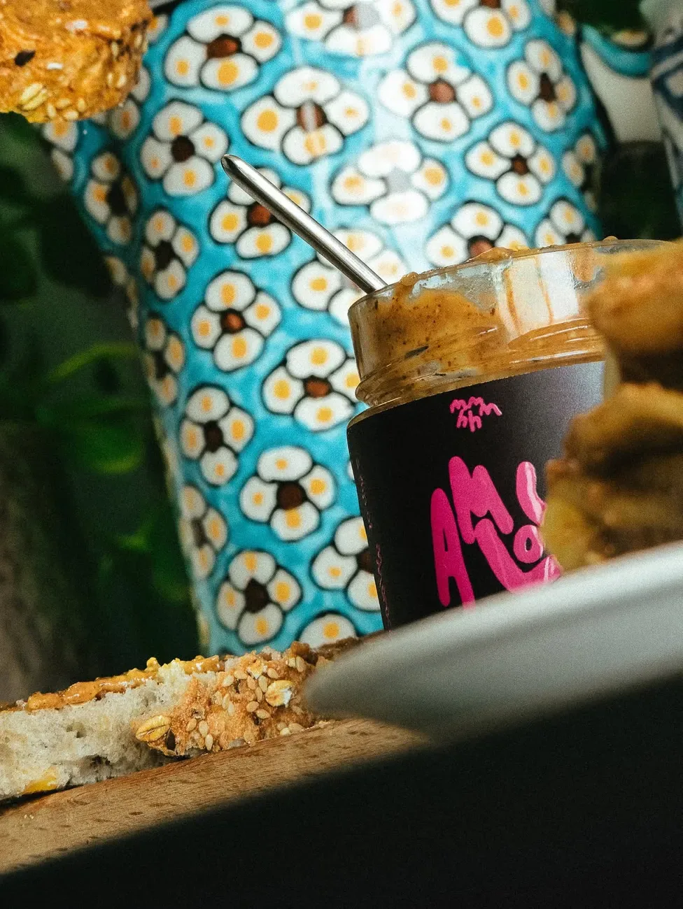 Open jar of MMMHH amlou with a spoon, next to toast spread with amlou, in front of a blue Moroccan ceramic vase